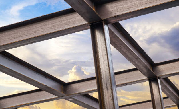 Steel beams form a building framework against a bright sky with clouds, showing intersecting metal structures and sunlight reflecting off the surfaces.