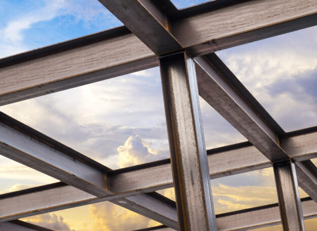 Steel beams form a building framework against a bright sky with clouds, showing intersecting metal structures and sunlight reflecting off the surfaces.