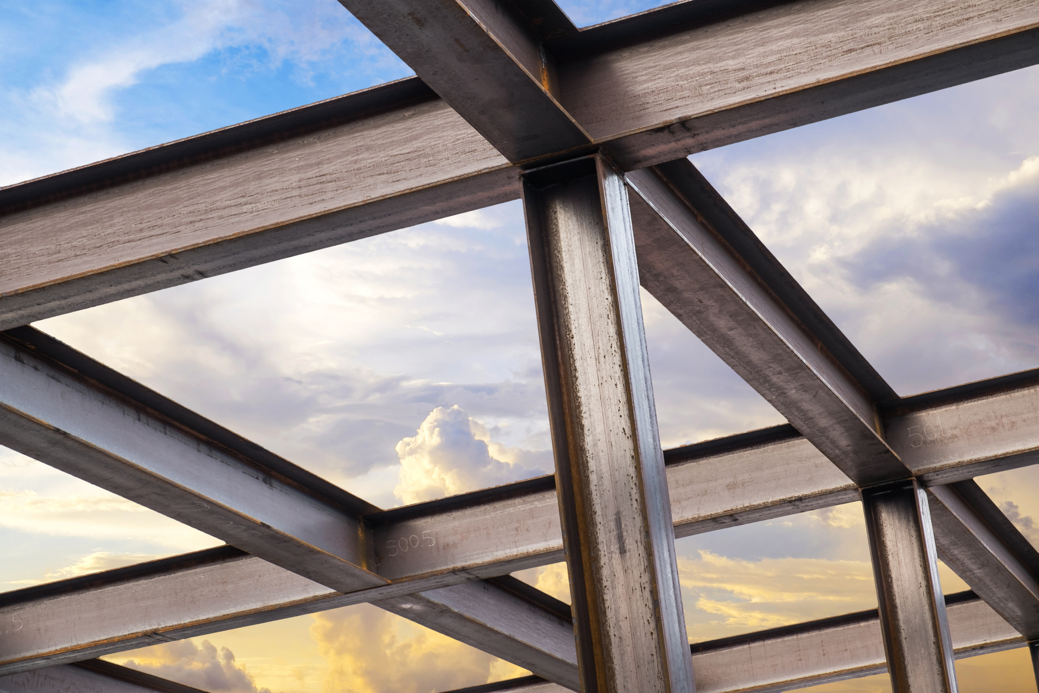 Steel beams form a building framework against a bright sky with clouds, showing intersecting metal structures and sunlight reflecting off the surfaces.
