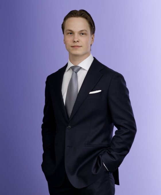 A young man in a dark suit and tie stands with hands in his pockets against a plain light purple background.