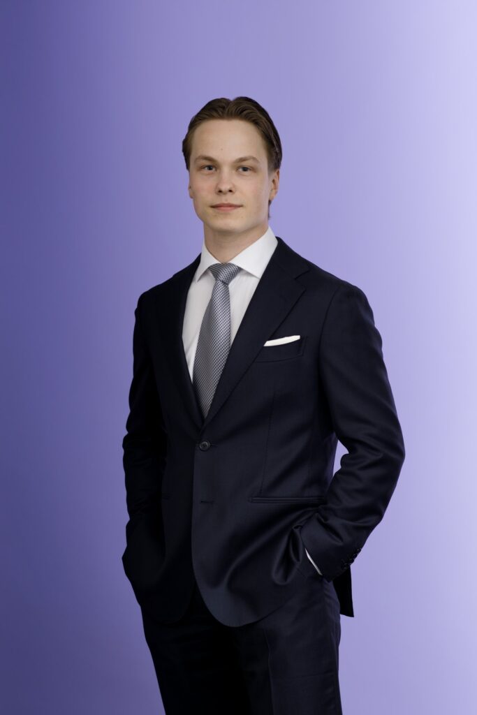 A young man in a dark suit and tie stands with hands in his pockets against a plain light purple background.