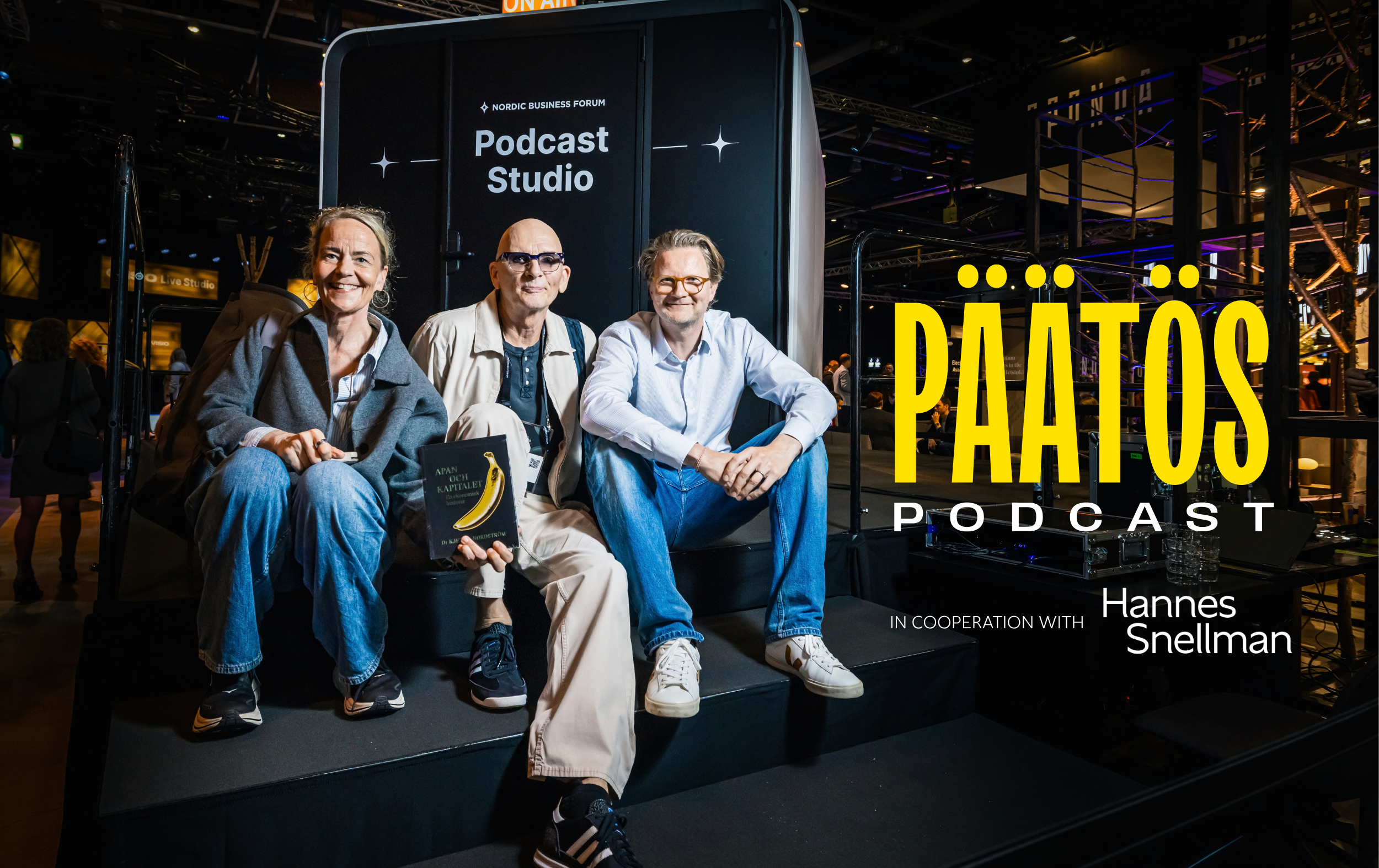 Three people sit smiling on steps outside a Podcast Studio booth. The word PÄÄTÖS PODCAST is prominent, along with in cooperation with Hannes Snellman. The setting appears modern with bright lighting.