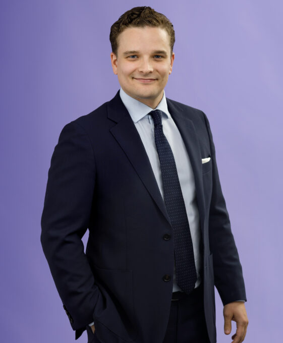 A man in a navy suit, white shirt, and dark tie stands against a plain light purple background, smiling with one hand in his pocket.