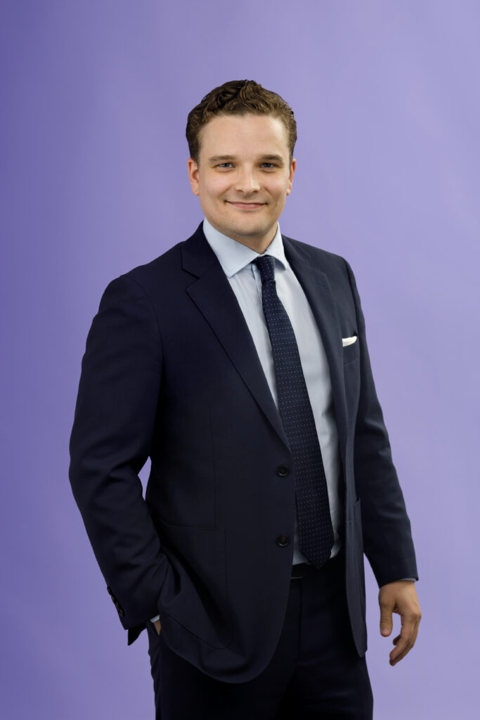 A man in a navy suit, white shirt, and dark tie stands against a plain light purple background, smiling with one hand in his pocket.