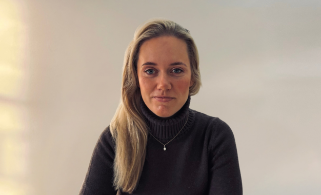 A woman with long blonde hair wearing a dark grey turtleneck sweater and a small pendant necklace.