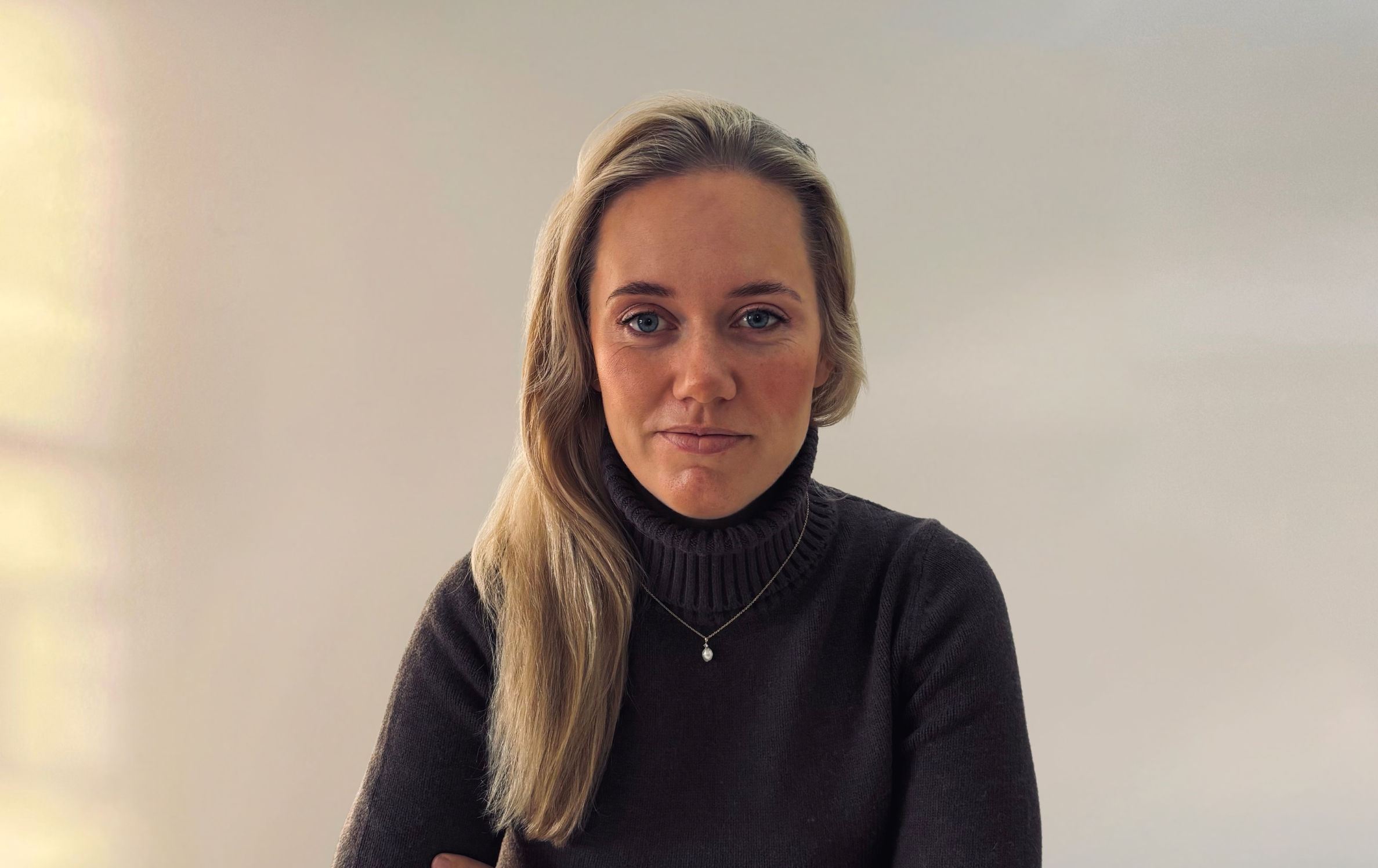 A woman with long blonde hair wearing a dark grey turtleneck sweater and a small pendant necklace.