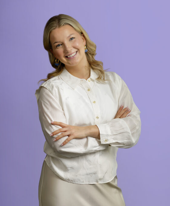 A smiling woman with blonde hair, wearing a white button-down shirt and blue earrings, stands with her arms crossed against a lavender background.