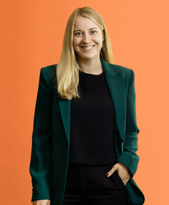 Smiling blonde woman wearing a dark green blazer over a black top against an orange background.