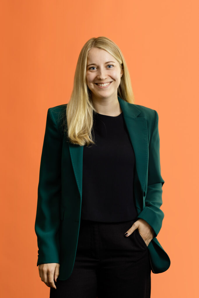 Smiling blonde woman wearing a dark green blazer over a black top against an orange background.