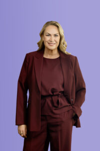A smiling blonde woman wearing a dark red blazer and matching blouse against a light purple background.