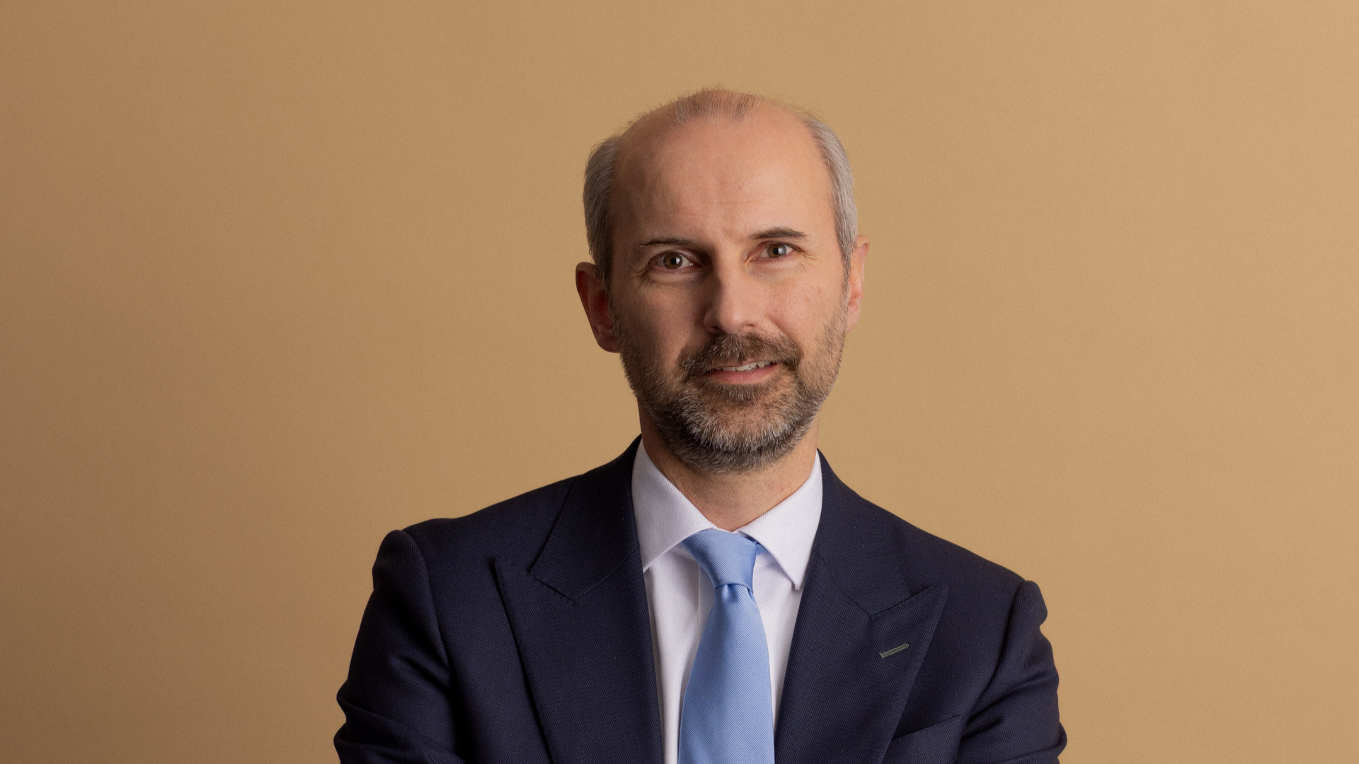 A middle-aged man with a beard wearing a dark suit, white shirt, and light blue tie against a solid tan background.